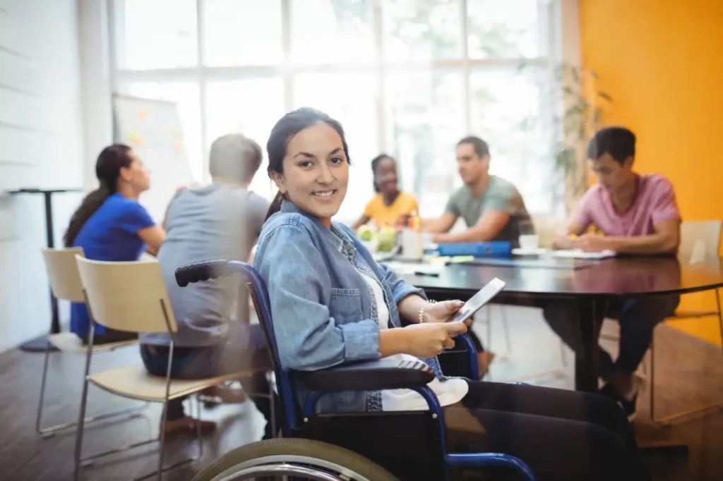 Smiling woman in wheelchair holding tablet at group table in Acals NDIS disabilities day program activity session with peers in Sydney