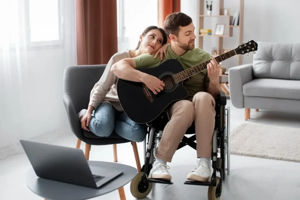 Man in wheelchair playing acoustic guitar with laptop nearby and companion beside him, taking Acals inclusive guitar lessons for NDIS participants