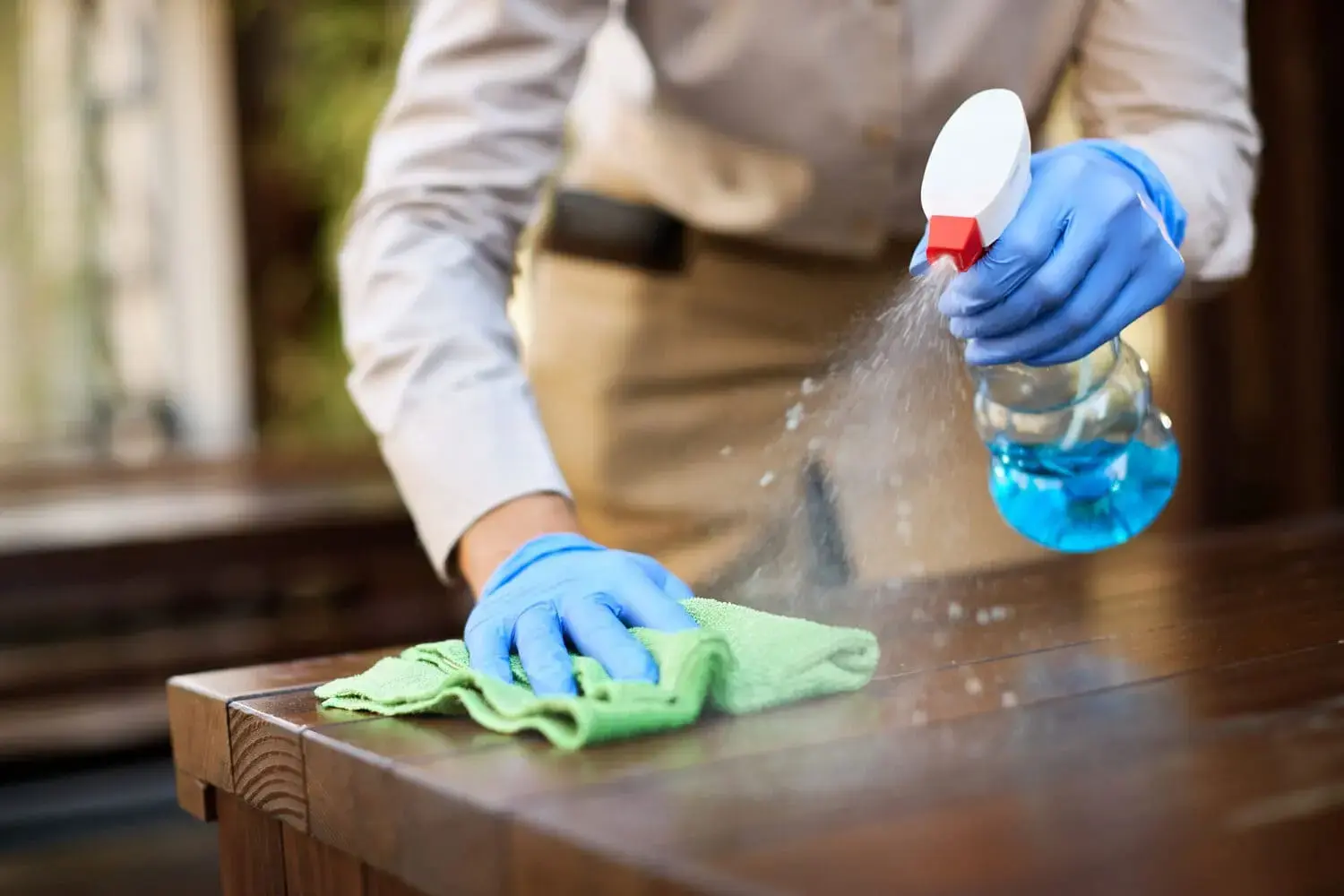 Cleaner with blue rubber gloves spraying cleaning solution and wiping wooden table surface for Acals NDIS domestic cleaning services Australia