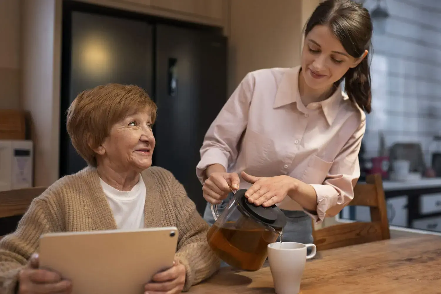 Young female carer pouring tea for elderly woman holding a tablet, representing Acals domestic assistance and aged care support services Australia