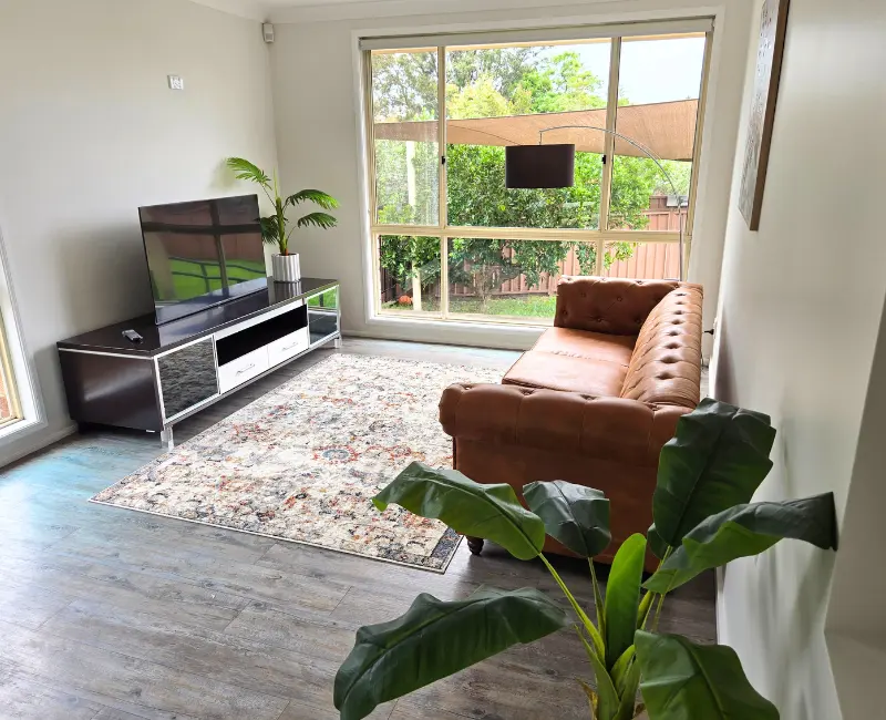 Cosy lounge room with flat screen TV, brown leather armchair, tropical plant and garden view at Acals NDIS accommodation